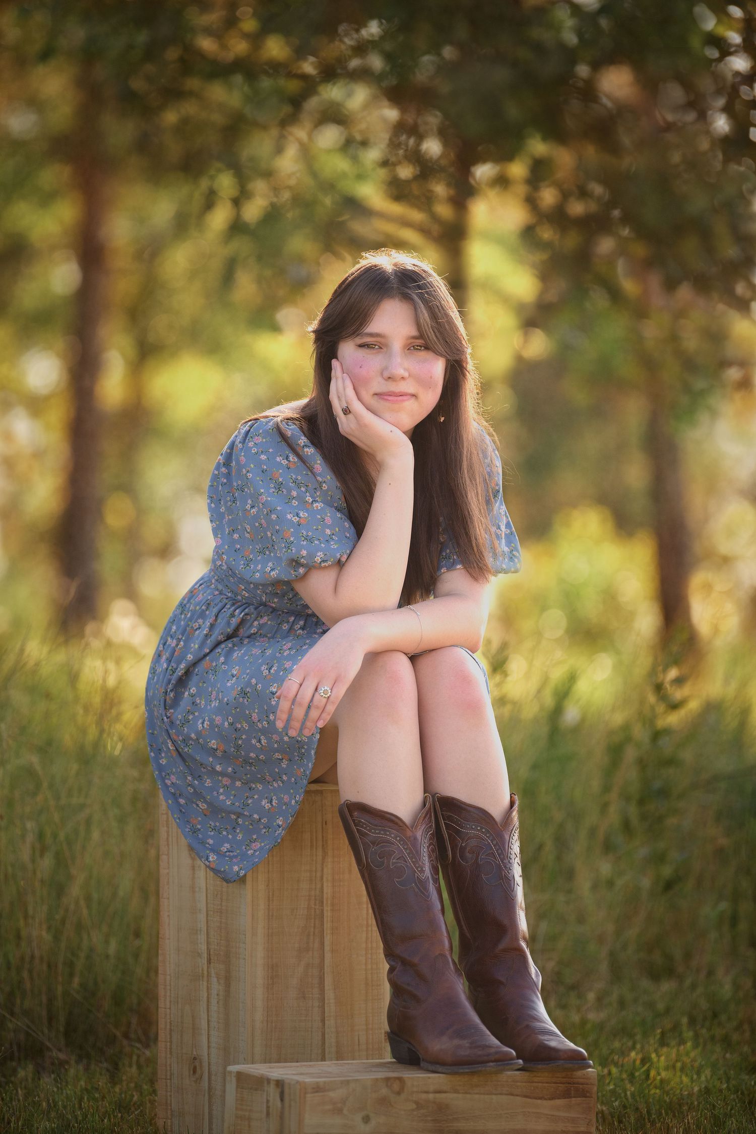 A girl in a blue floral dress and brown cowboy boots sits on a wooden platform in a sunlit field.