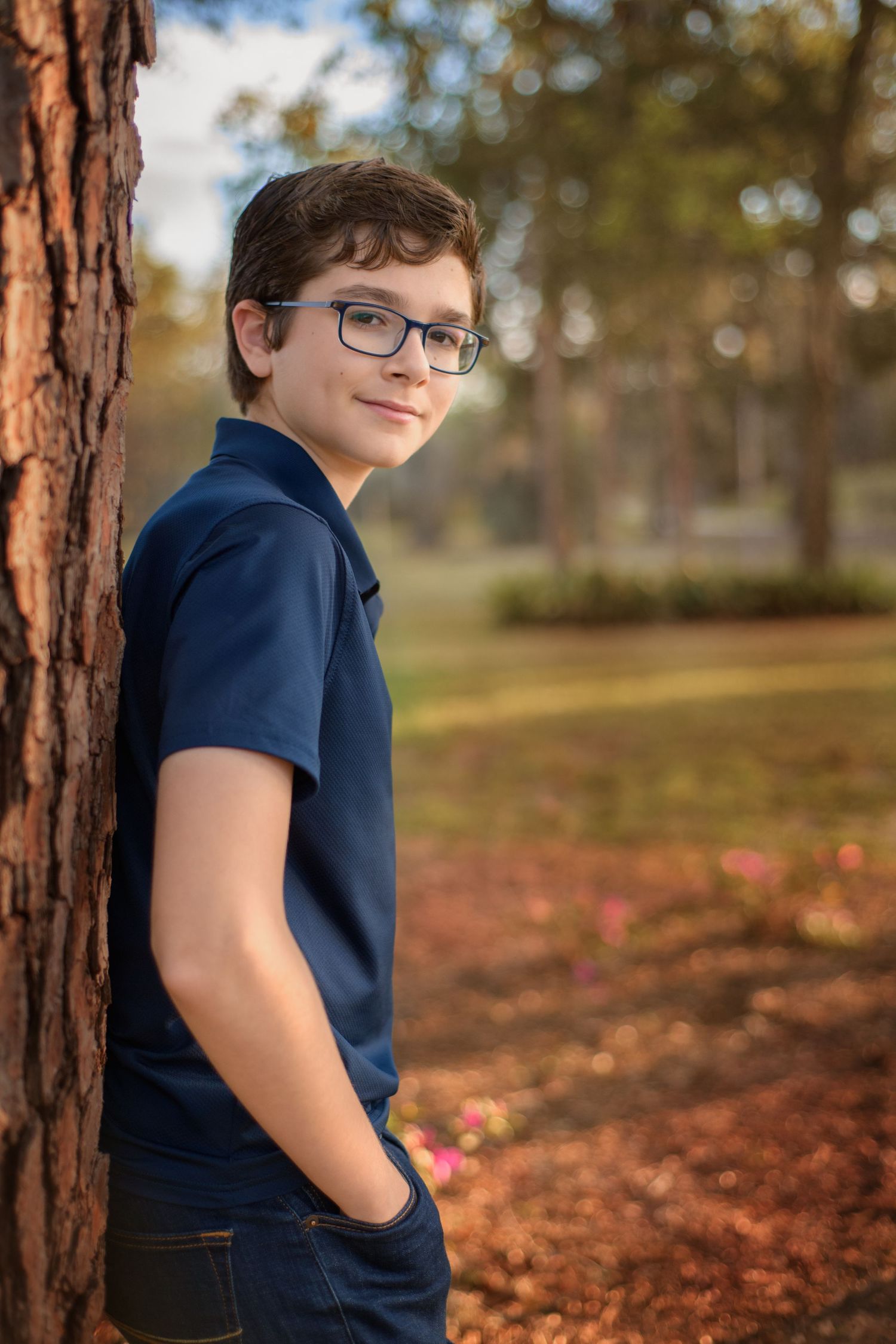 Casual portrait of senior teenager leaning against pine tree in autumn park setting.