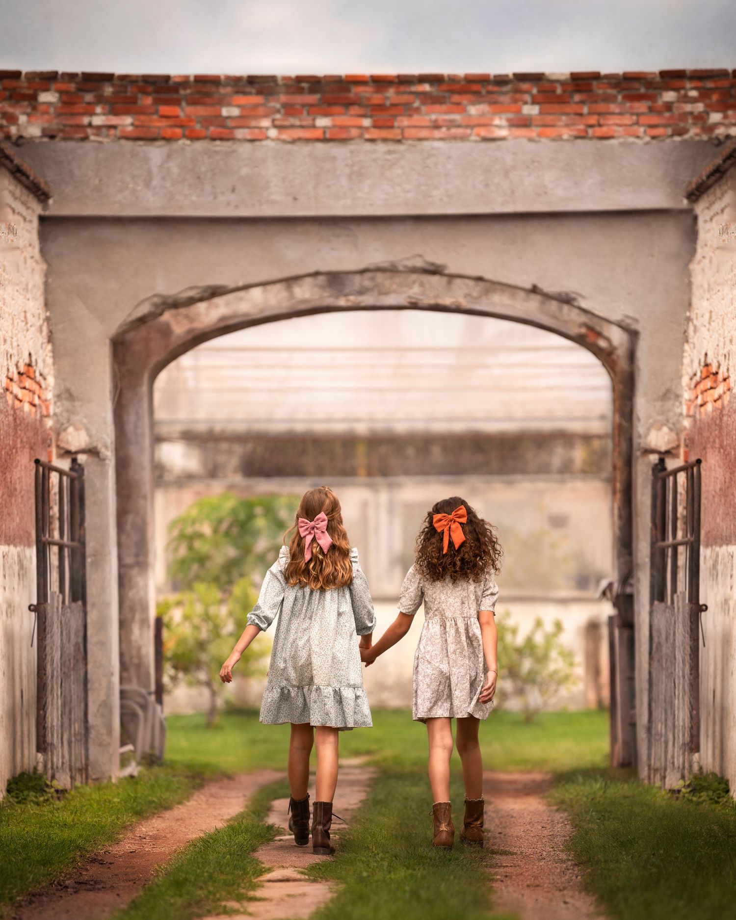 Two children in white dresses walk together through an arched tunnel with brick walls and sunlit path ahead.