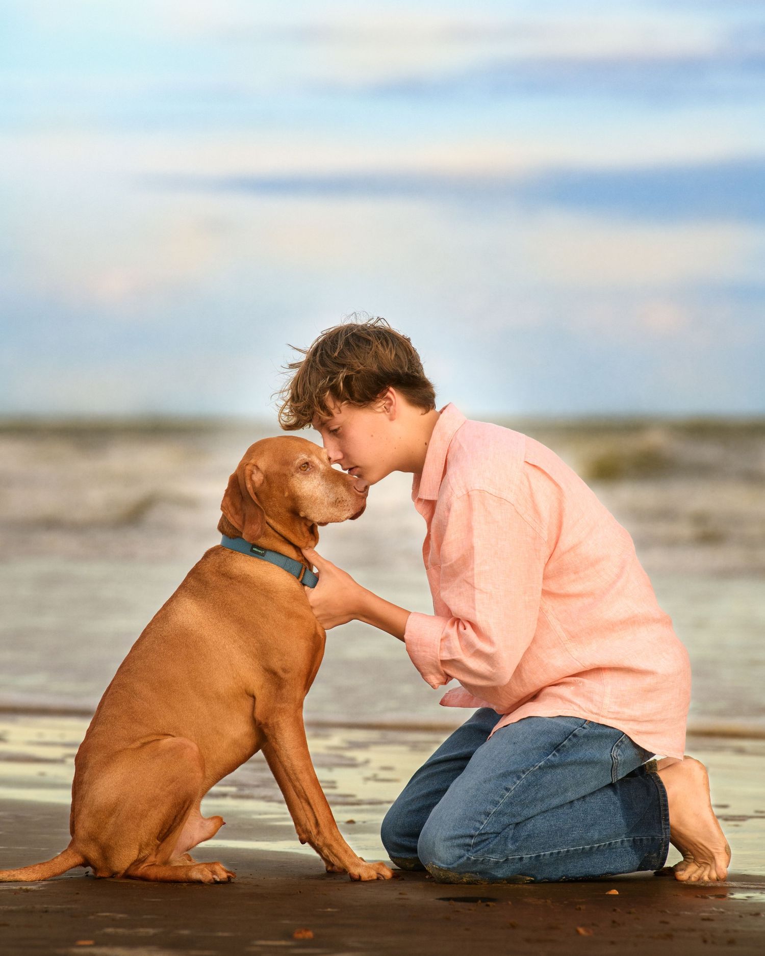 Teenager in pink sweater sharing affectionate moment with brown dog on beach.