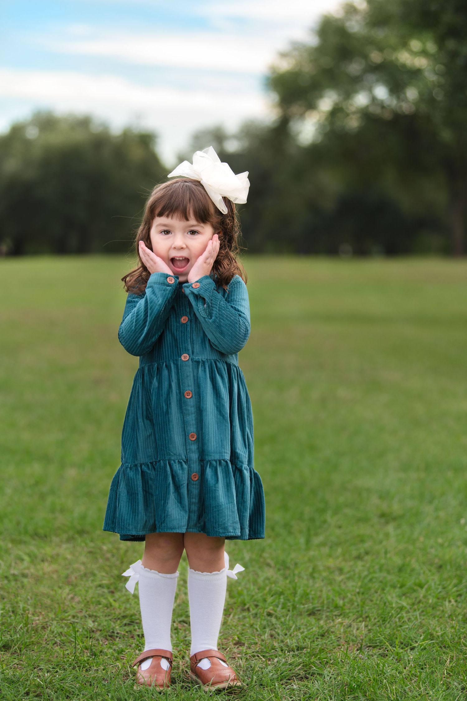 Young child in teal dress and white knee socks stands on grass with hands on cheeks in playful pose.