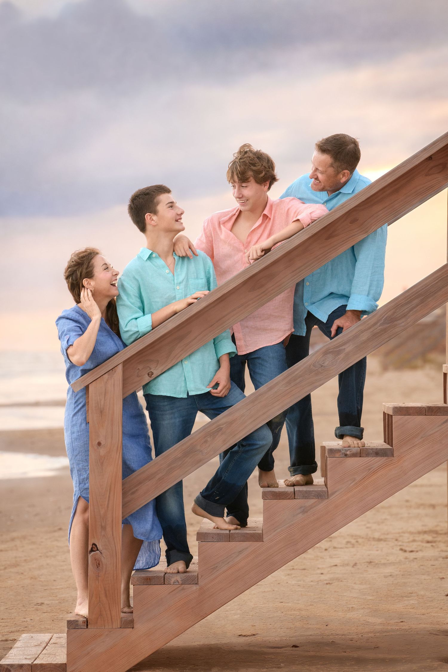 Family members interact playfully on wooden stairs at sunset on a sandy beach.
