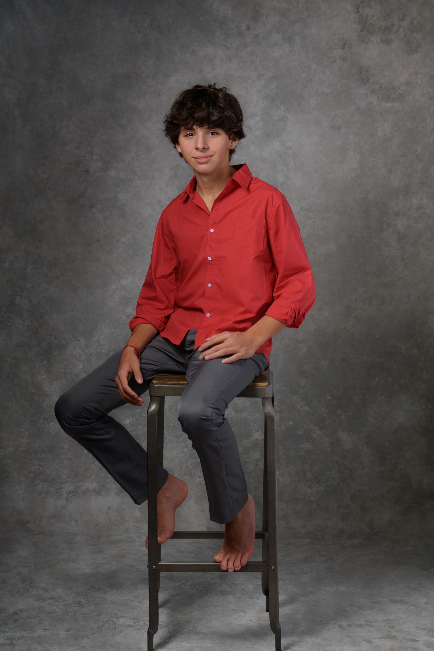 Teenager in red button-down shirt sitting casually on wooden stool against grey backdrop.