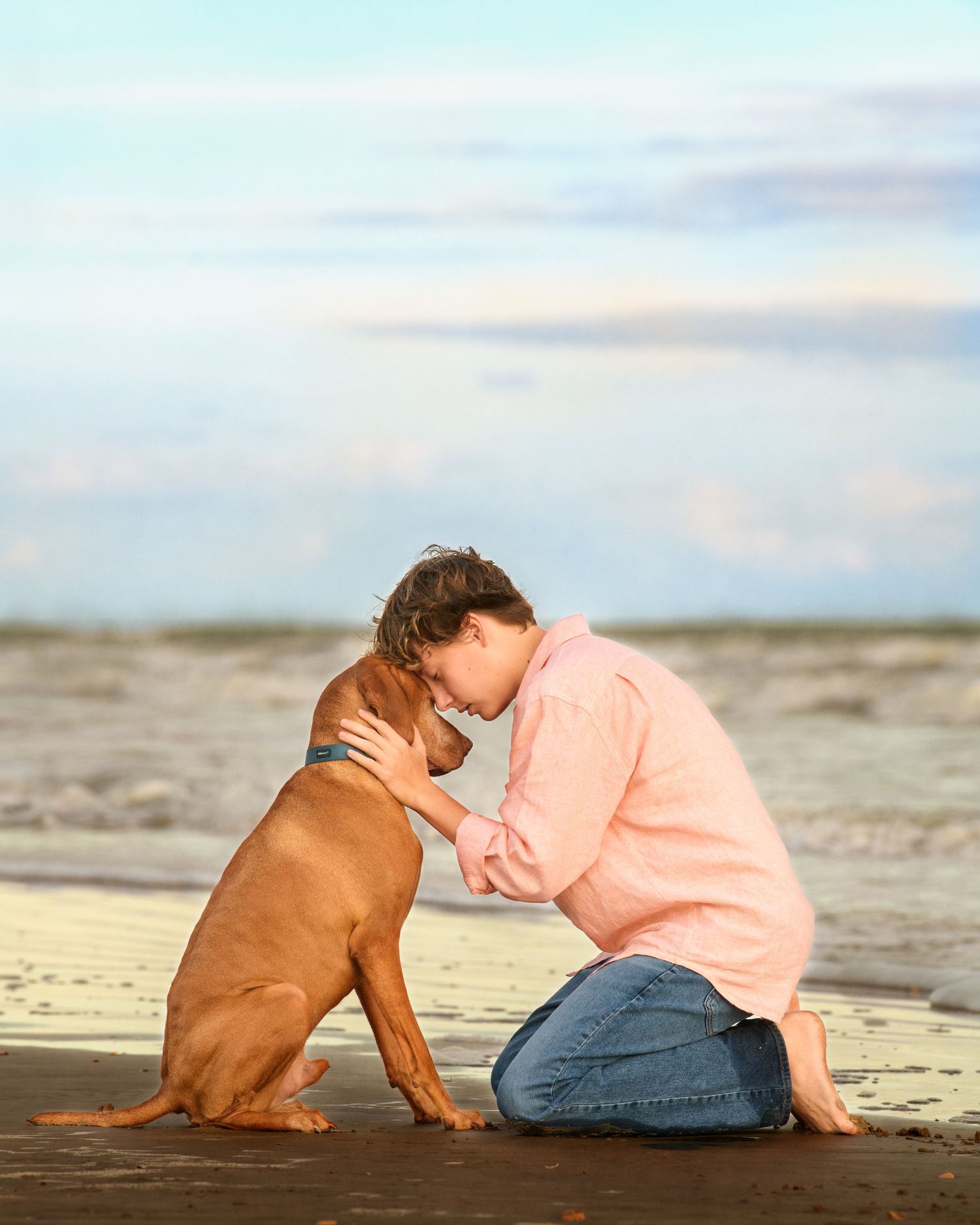 Teenager in pink sweater sharing tender moment with brown dog on beach boardwalk.