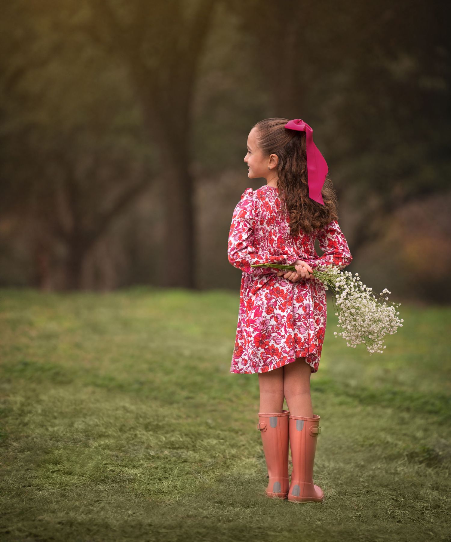 Child wearing pink floral dress and rain boots holds wildflowers in a green field.
