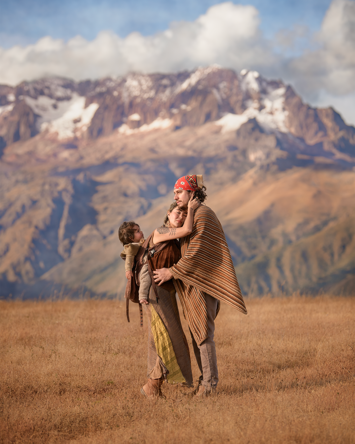 Two people wearing warm clothing embrace while standing in a golden field with snow-capped mountains in the background.