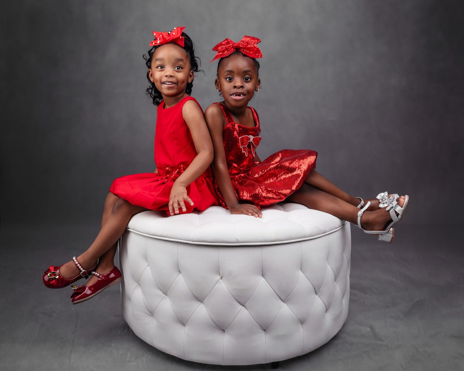 Two children in matching red outfits pose on white tufted ottoman.