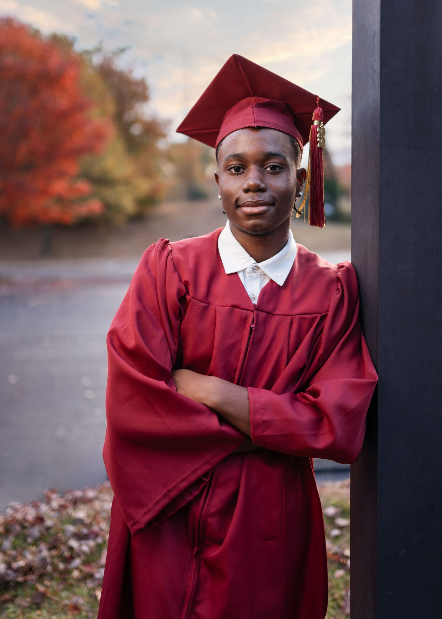 Graduate in burgundy cap and gown poses outdoors during fall season.
