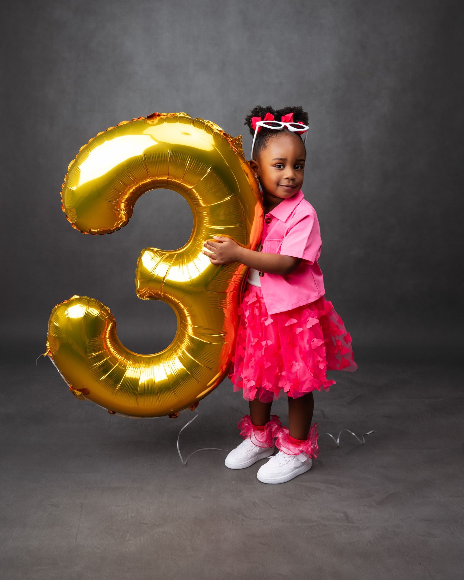 Child posing with large gold number 3 balloon in pink dress against dark studio background.