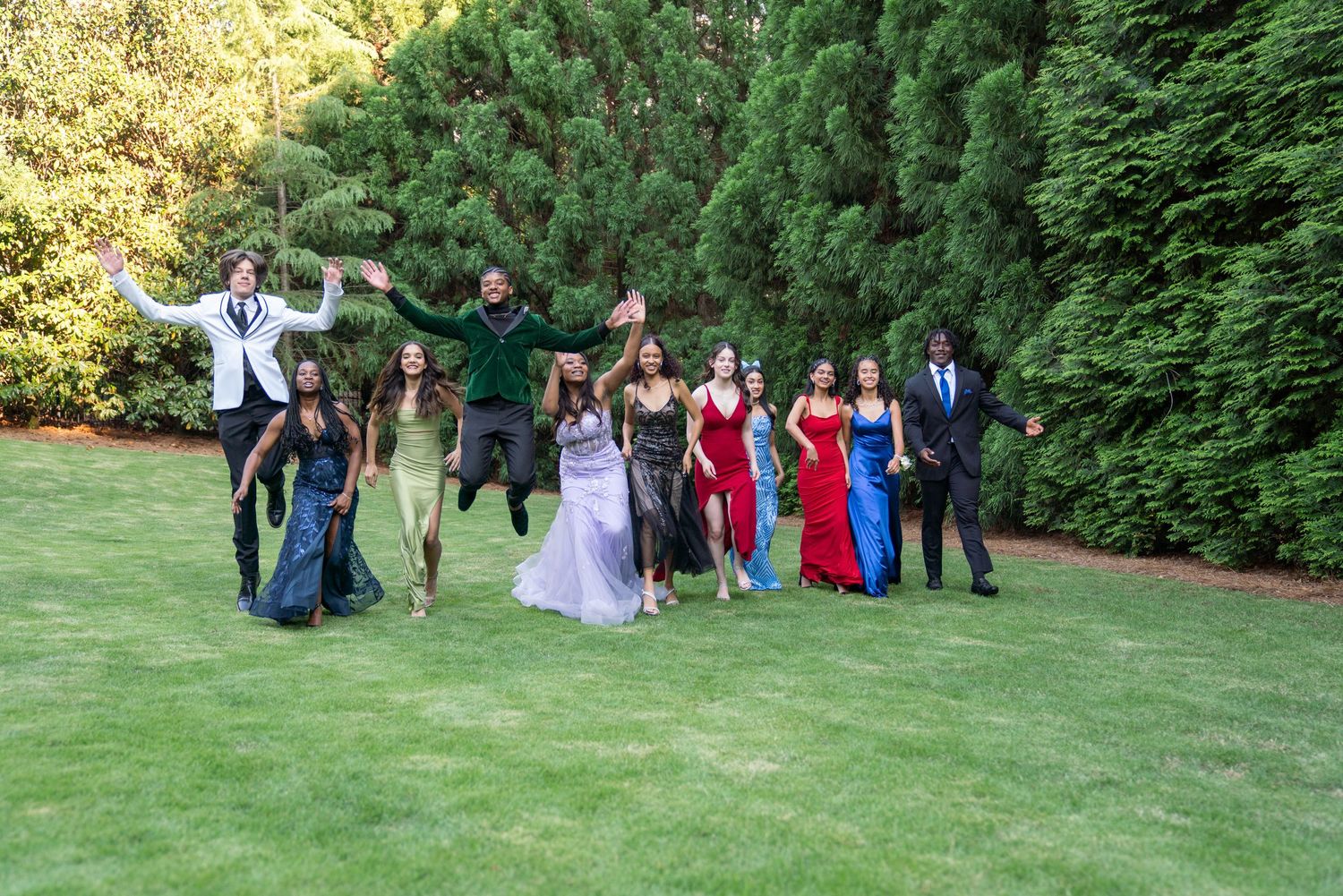 Wide group shot of formal event attendees gathered on grass with evergreen trees in background.