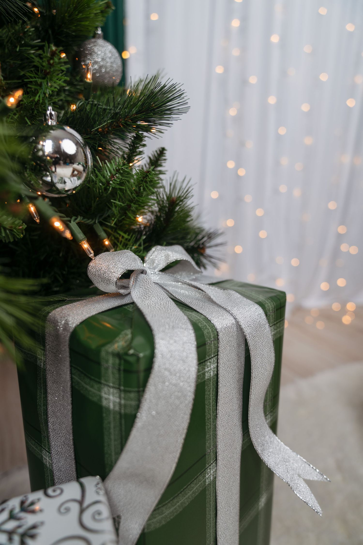 Green plaid Christmas gift with a sparkly silver ribbon bow beside a decorated tree with silver ornaments and soft twinkling lights.