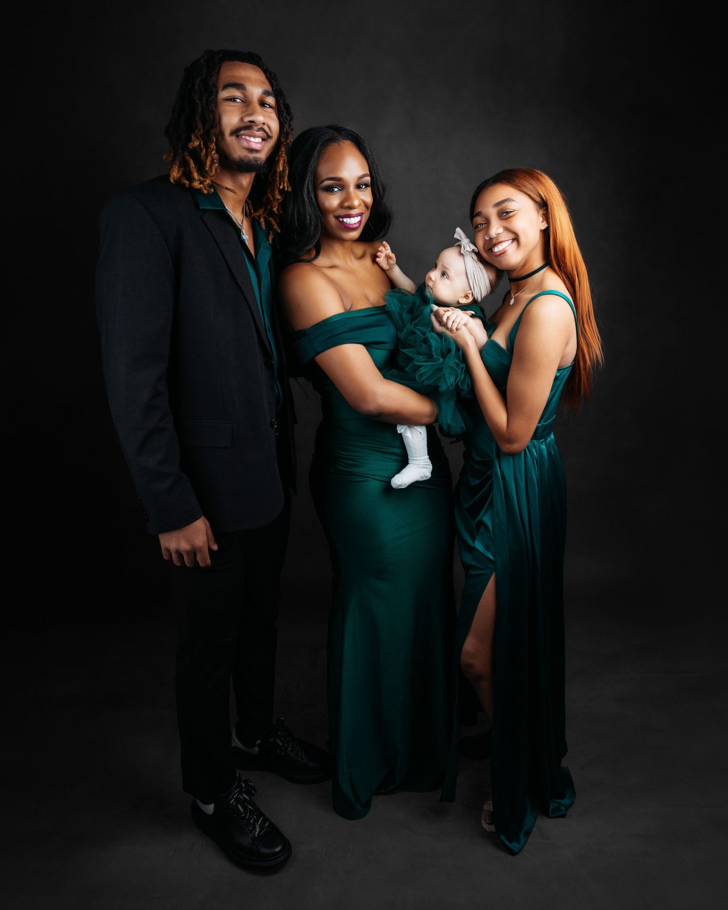 Family of three adults in emerald green and black formal wear pose together holding a newborn baby against a dark backdrop.