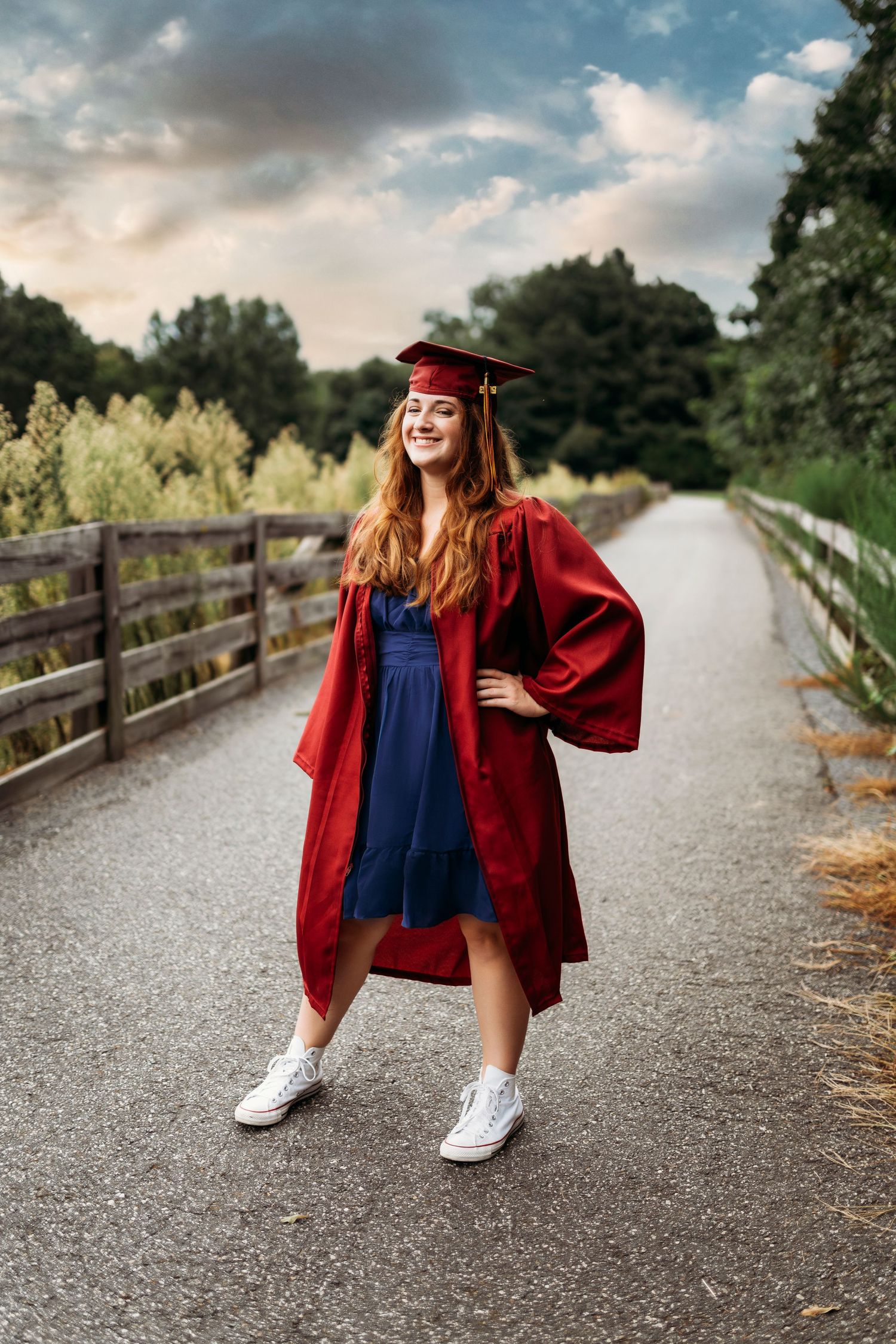 Graduate in red cap and gown poses playfully on path with white sneakers.