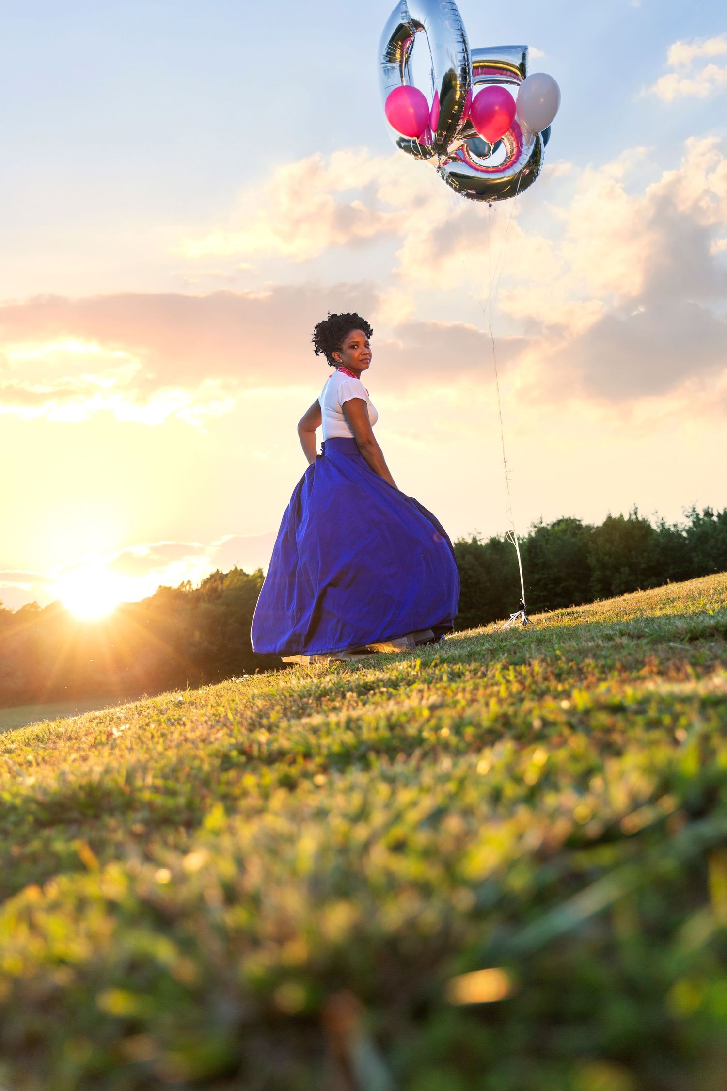 Person in flowing blue dress twirling with balloons in a sunset meadow.