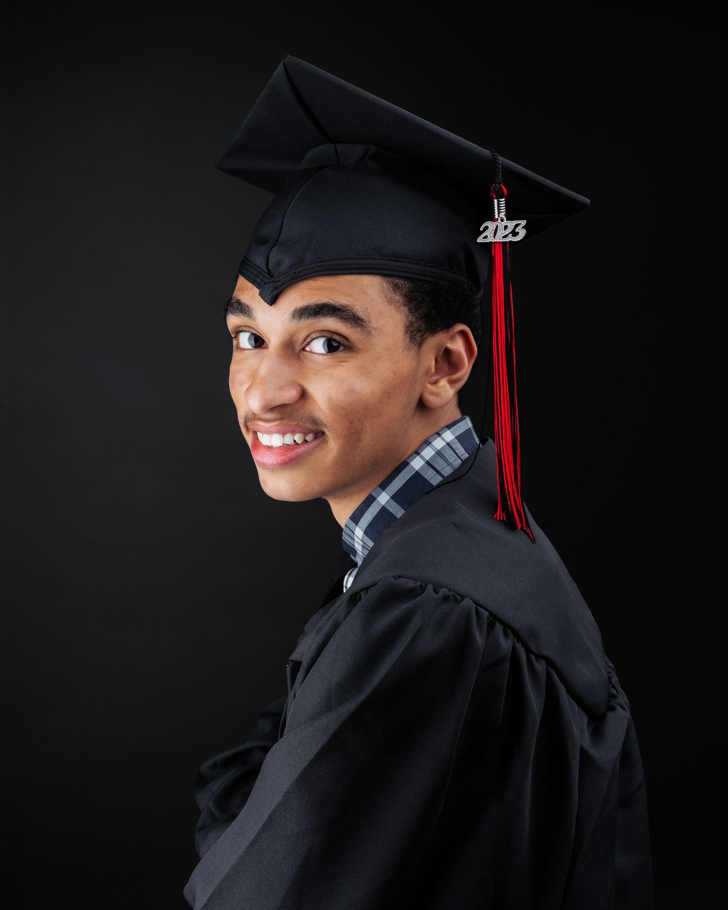 Graduate in black cap and gown poses confidently against dark backdrop.