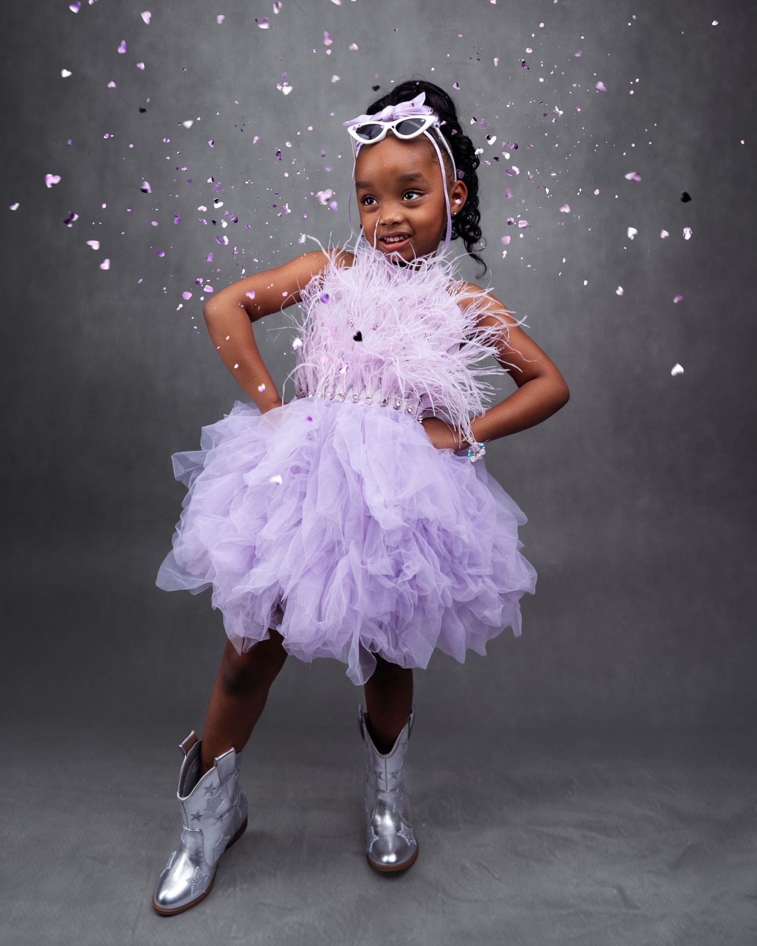 Child in lavender tulle dress and silver boots poses playfully with falling confetti.