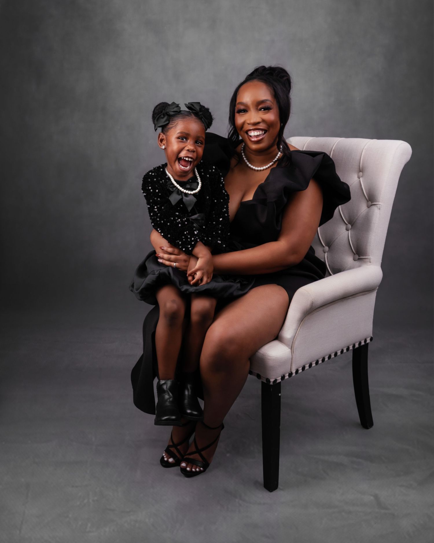 Elegant portrait of parent and child in black formal wear seated in gray chair.