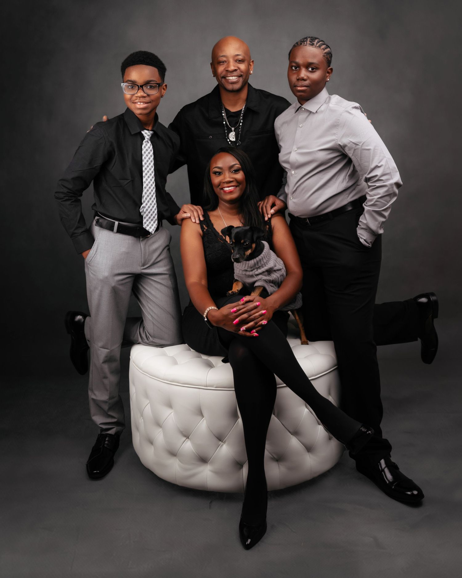 Professional family studio portrait on tufted ottoman against grey backdrop.
