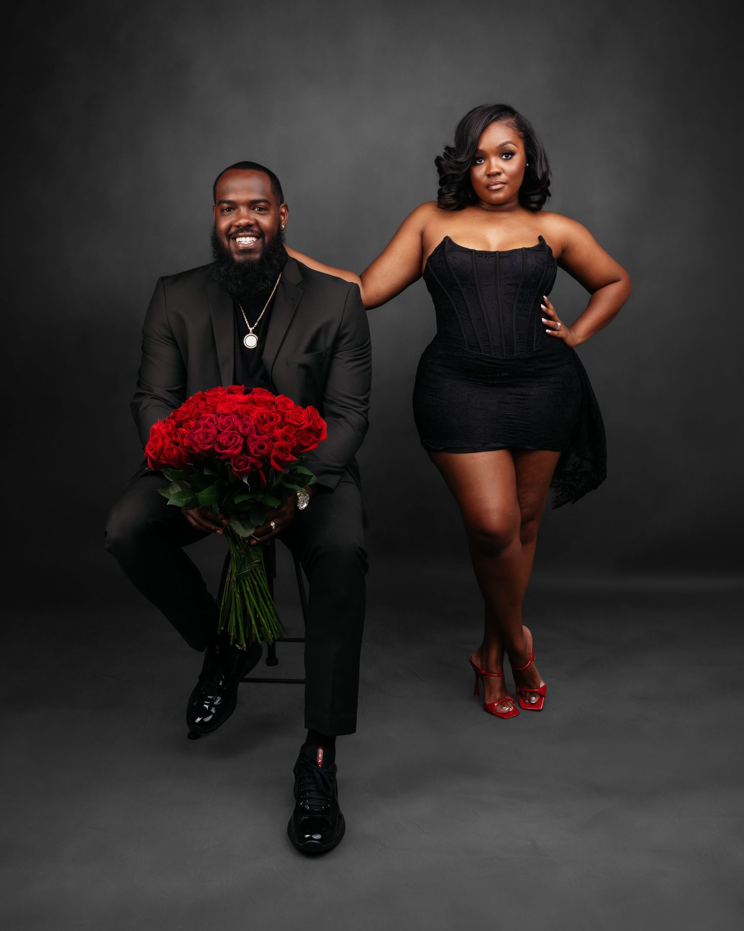 Romantic couple's photoshoot with red roses and formal attire against dark studio background.