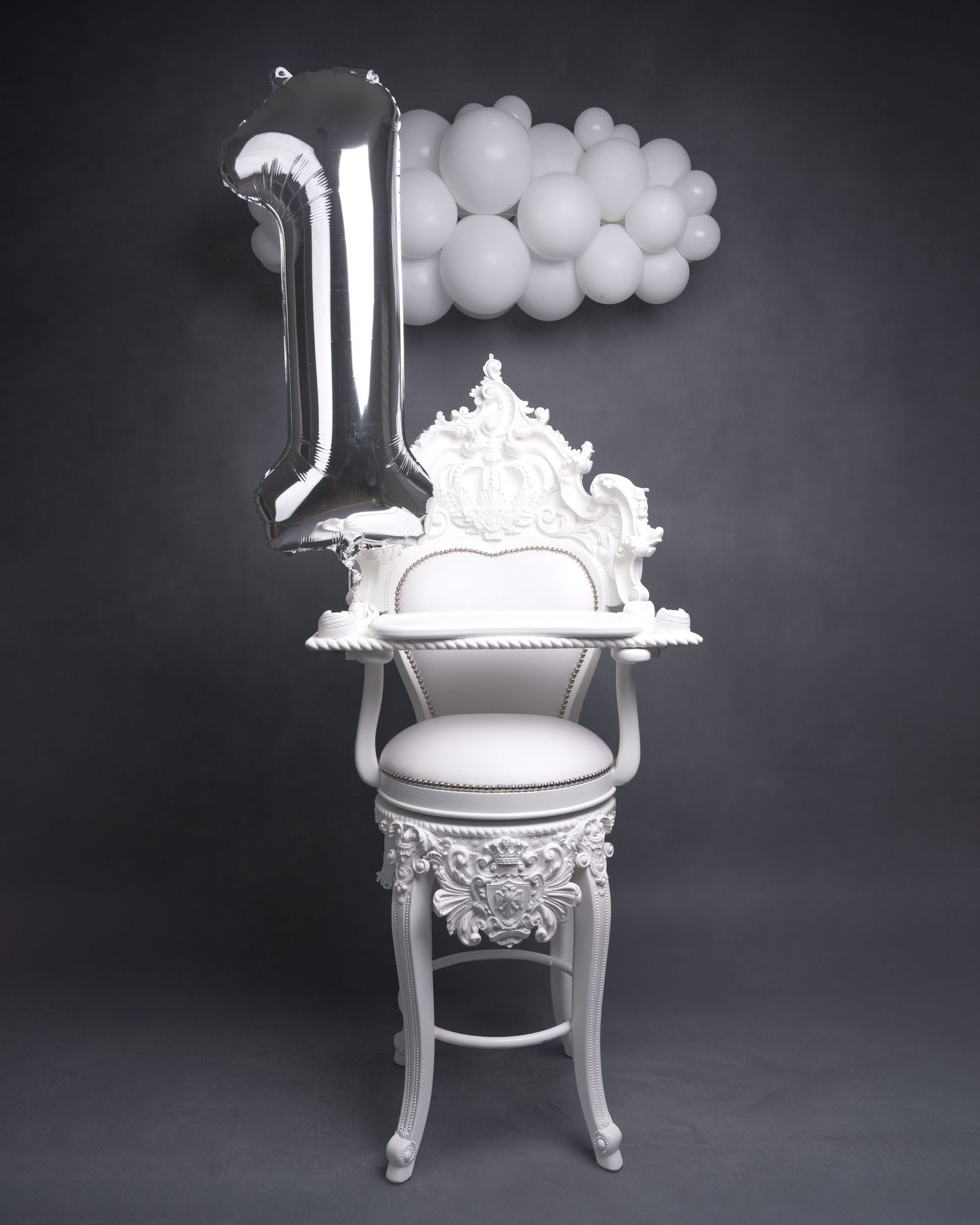 An ornate white baroque-style chair decorated with balloons and a silver number one balloon.