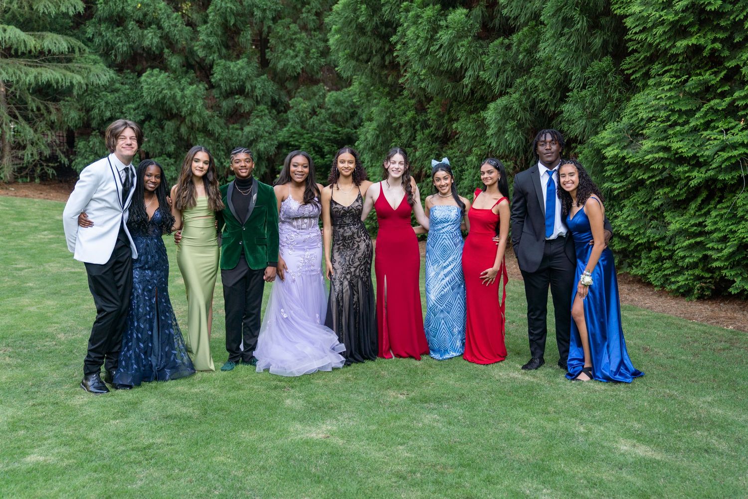 Large group photo of formal event attendees in colorful dresses and suits posing outdoors on grass near trees.