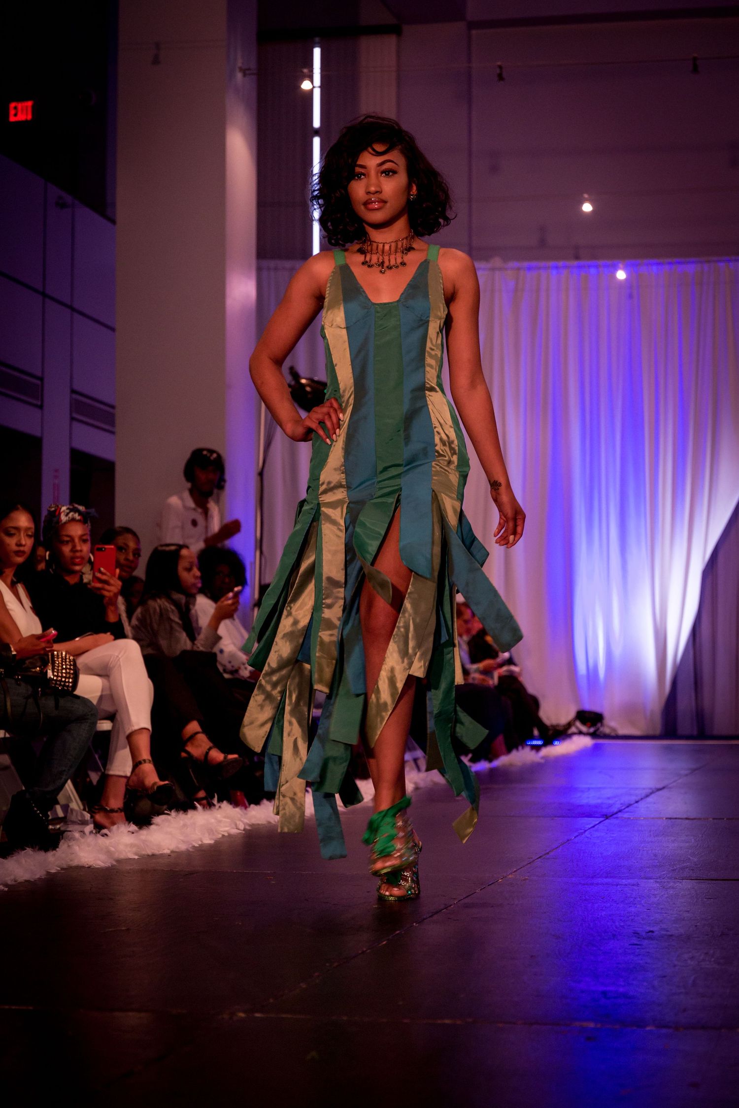 Model walks runway in flowing teal dress during fashion show with dramatic blue lighting and white curtain backdrop.