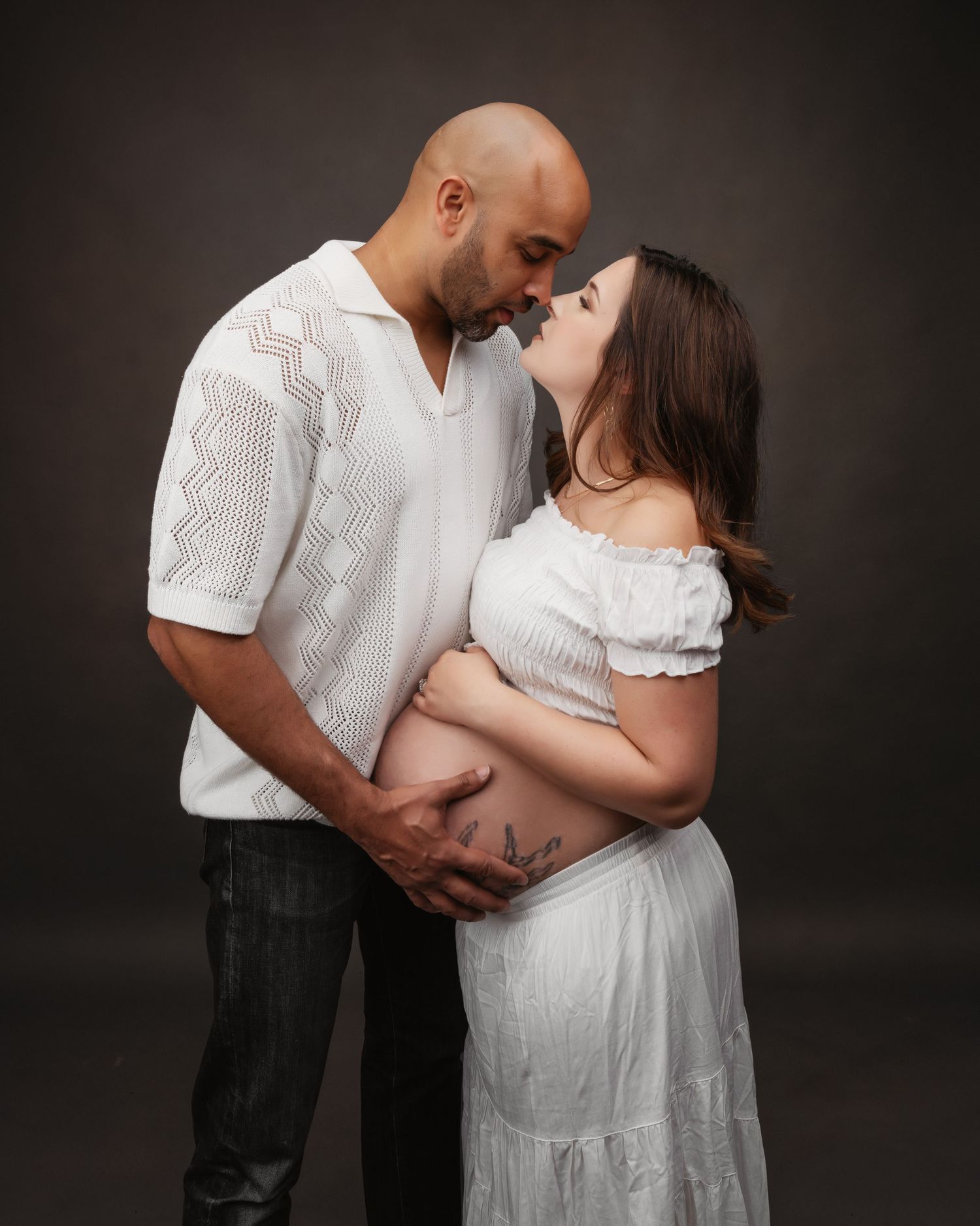 Maternity photoshoot with couple in white outfits showing tender moments against dark backdrop.