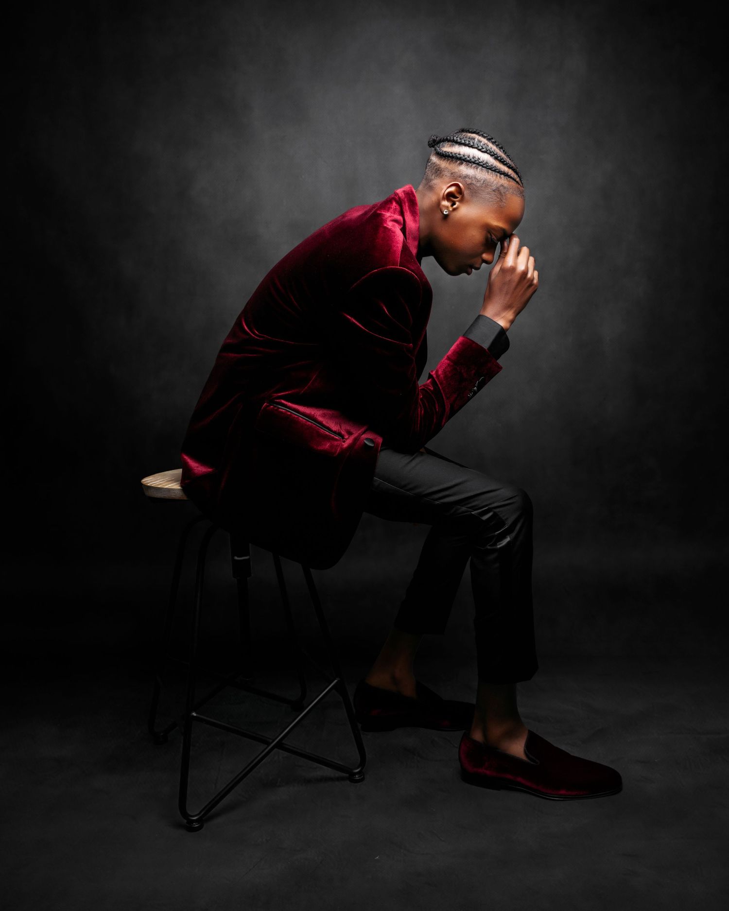 Person in burgundy velvet outfit sitting thoughtfully on chair against dark backdrop.
