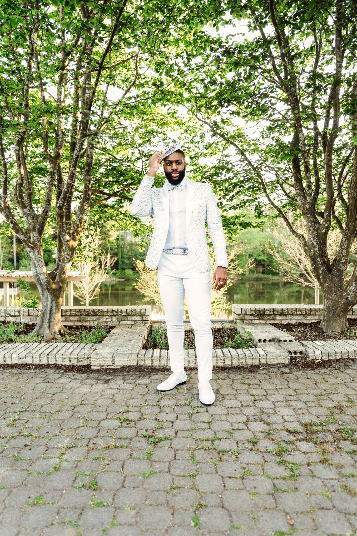 Person in white formal attire poses on stone paved courtyard surrounded by green trees.