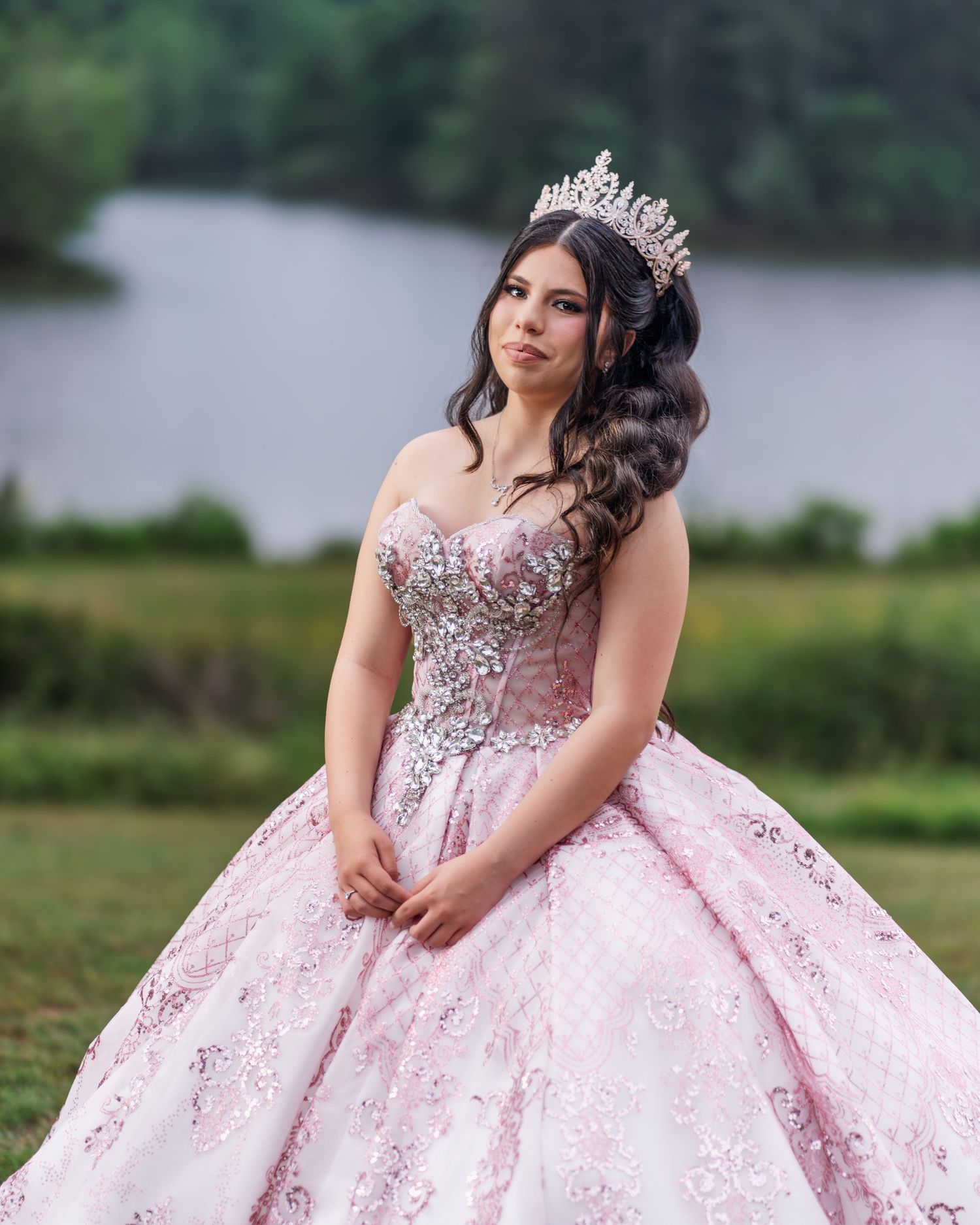 A stunning pink quinceañera ball gown with crystal embellishments and tiara photographed by a lake.