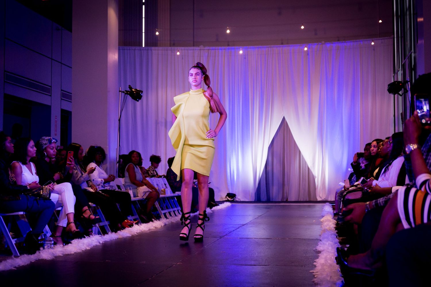 Fashion show venue with purple lighting and white backdrop showing runway and audience seating.