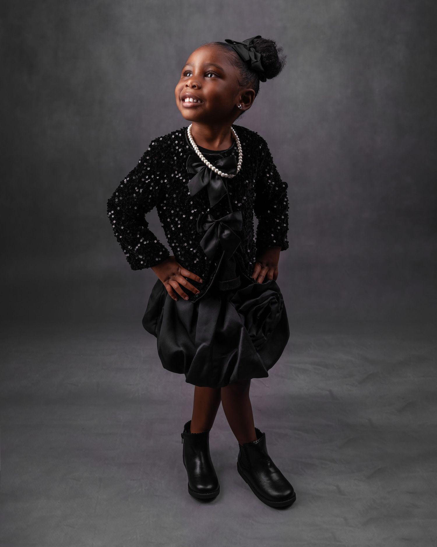 A young child poses confidently in a sparkly black dress and boots against a dark studio backdrop.