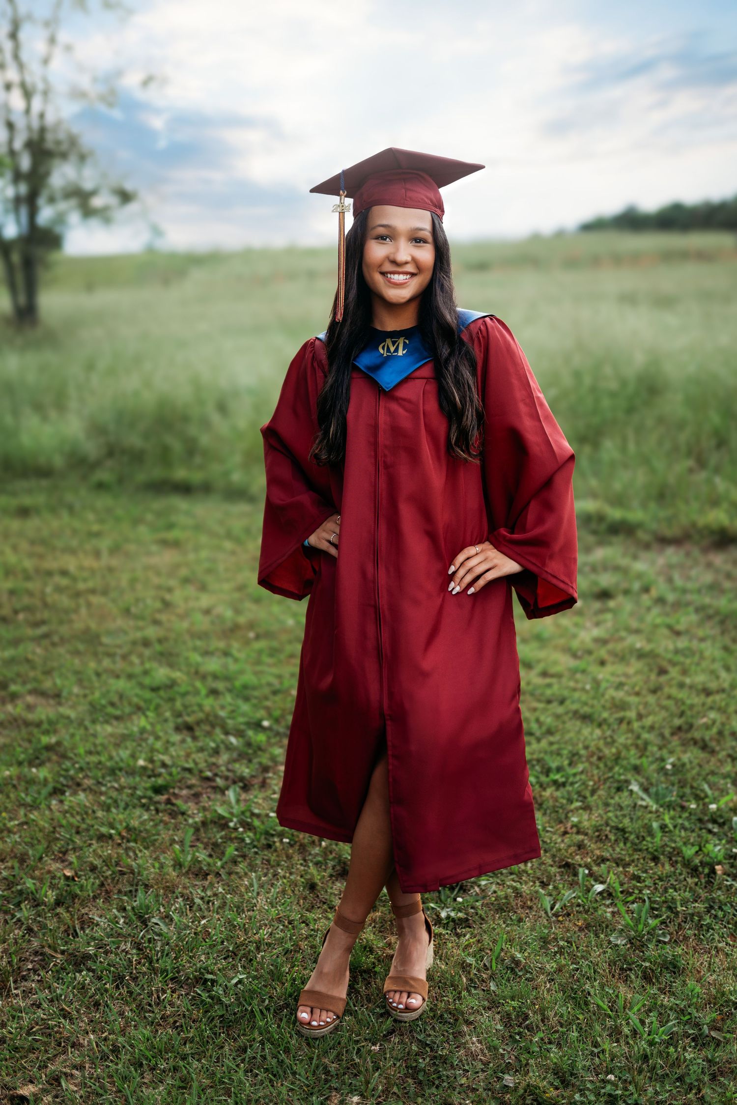 A graduate poses in traditional red cap and gown on a grassy hillside.