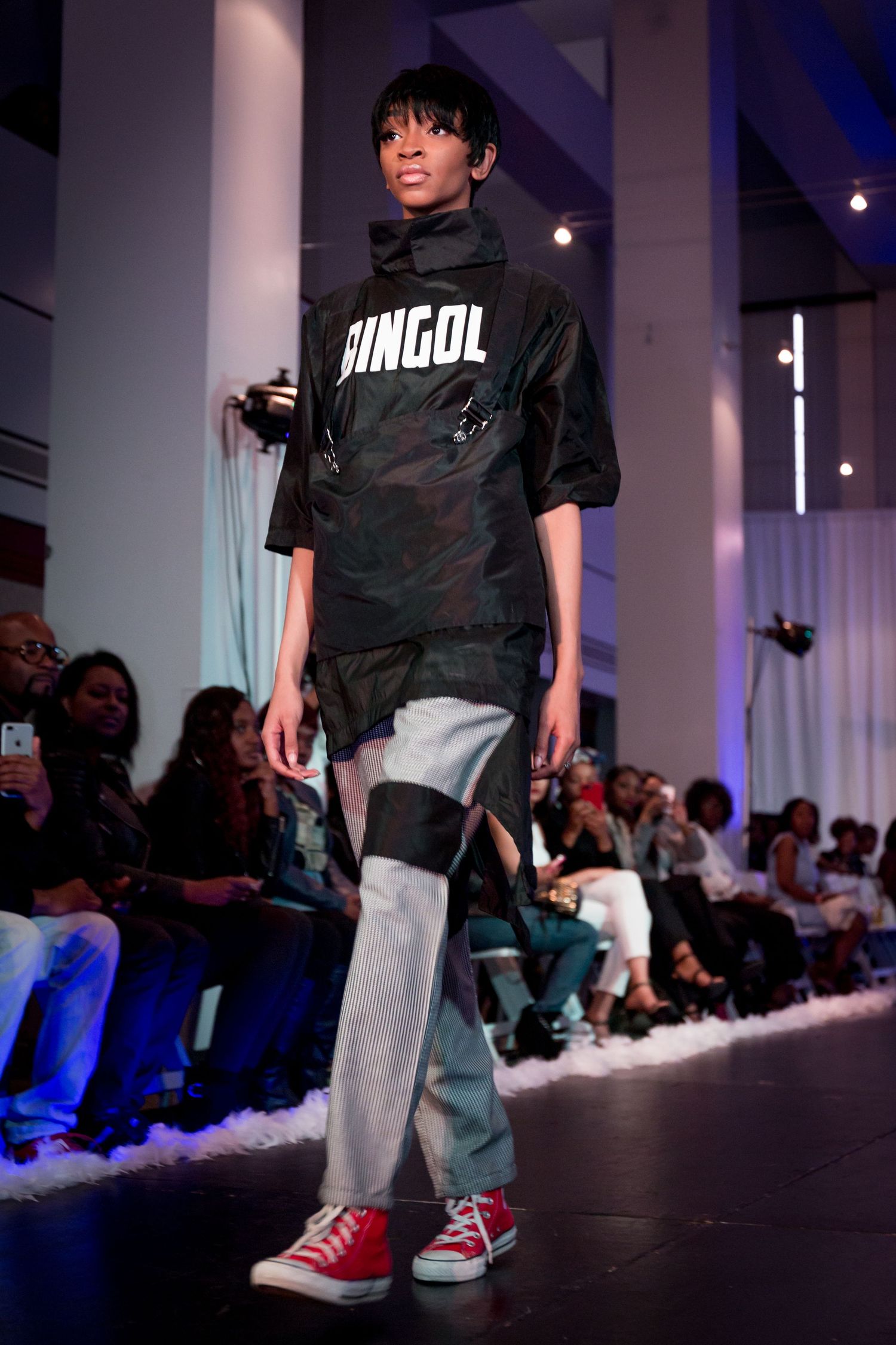Streetwear fashion model showcases black DINGOL t-shirt with gray pants and red sneakers on illuminated runway.