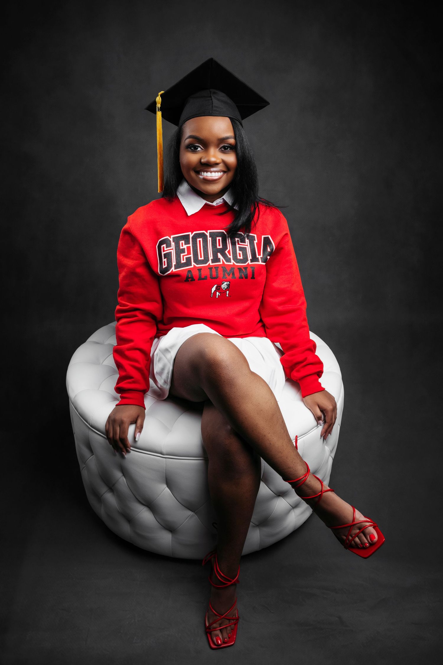 Graduate in red Georgia sweatshirt poses on white chair against dark backdrop for professional portraits.