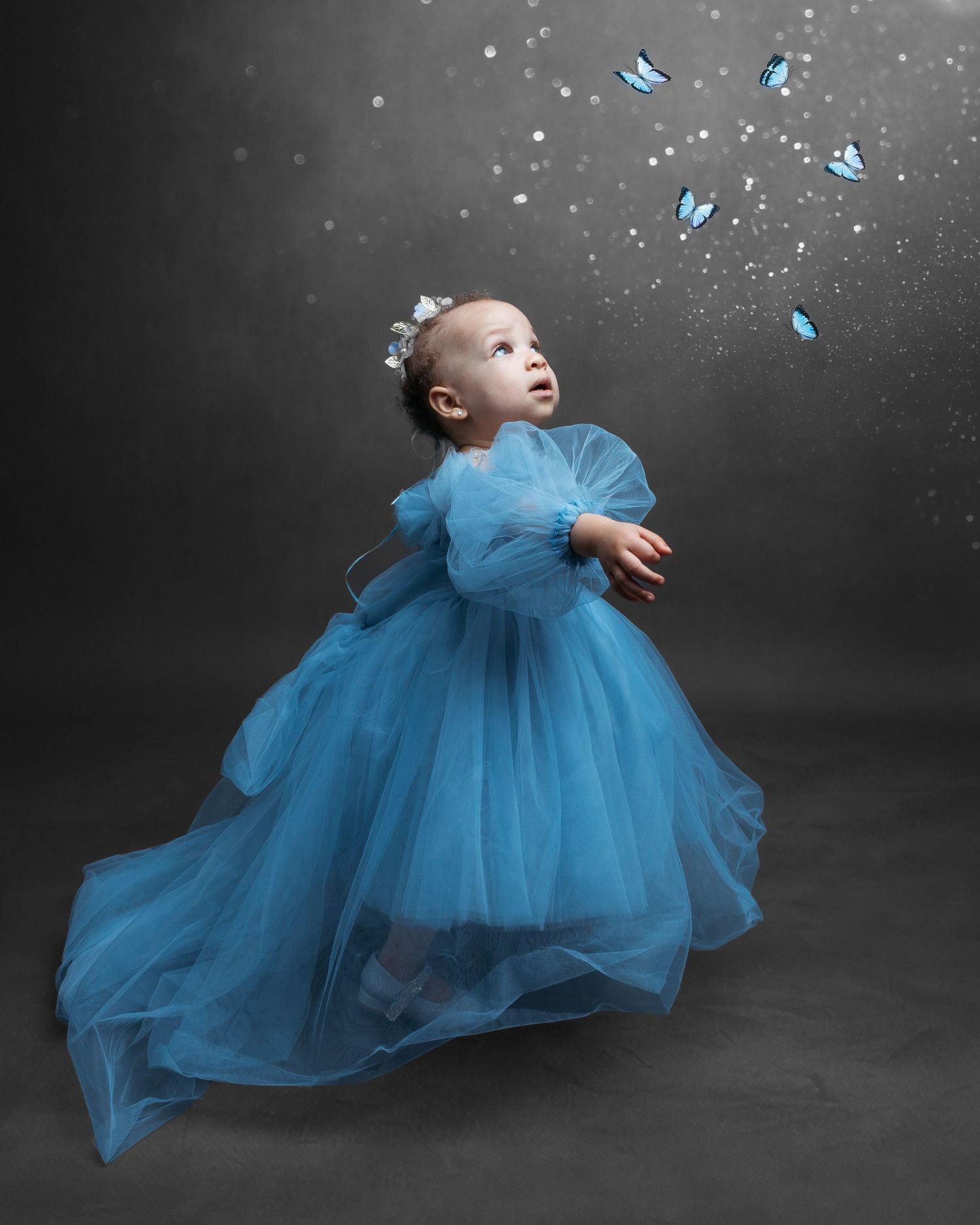 Baby in flowing blue tulle dress with butterfly accents against grey backdrop.