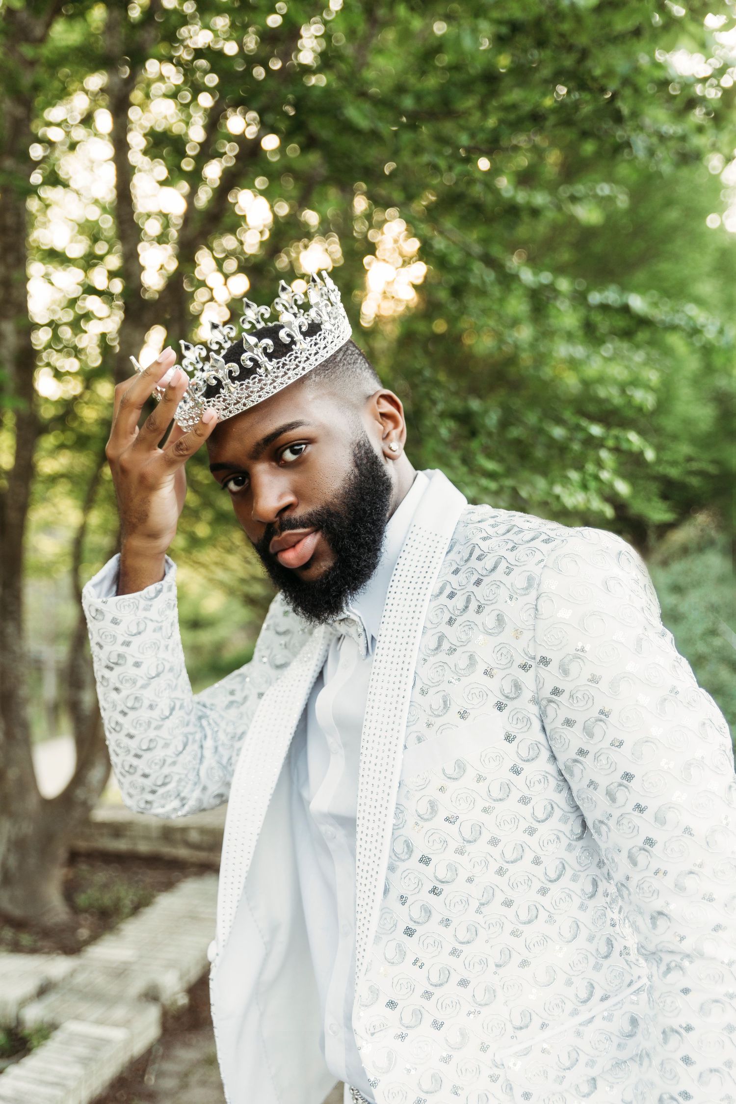Person wearing ornate white lace outfit and crown poses in outdoor garden setting.