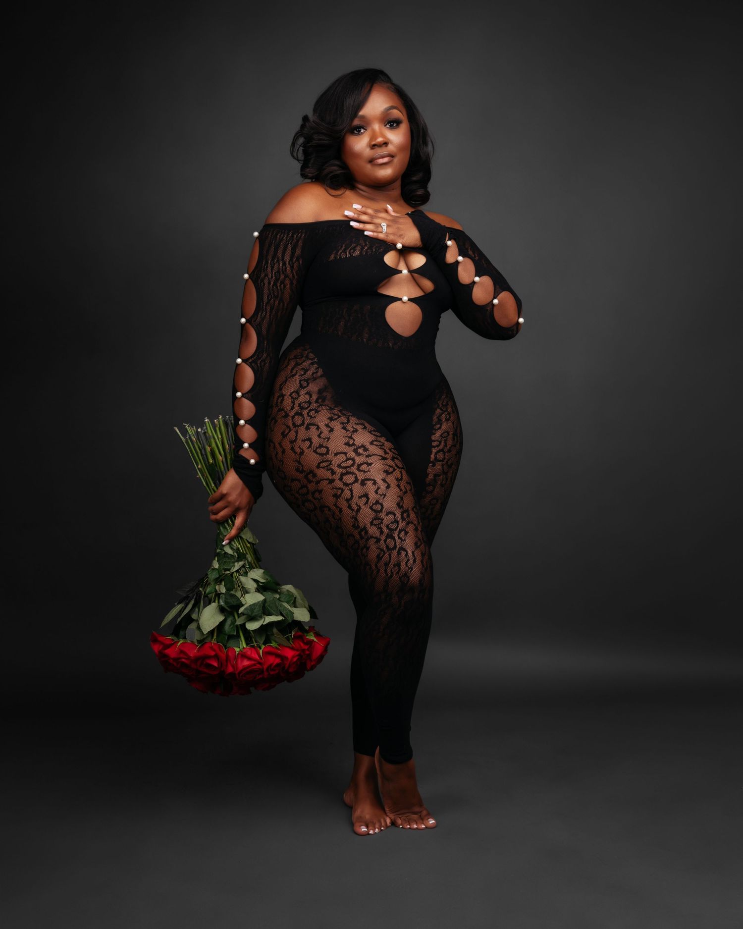 Elegant figure in a black lace dress posing with red roses against dark backdrop.