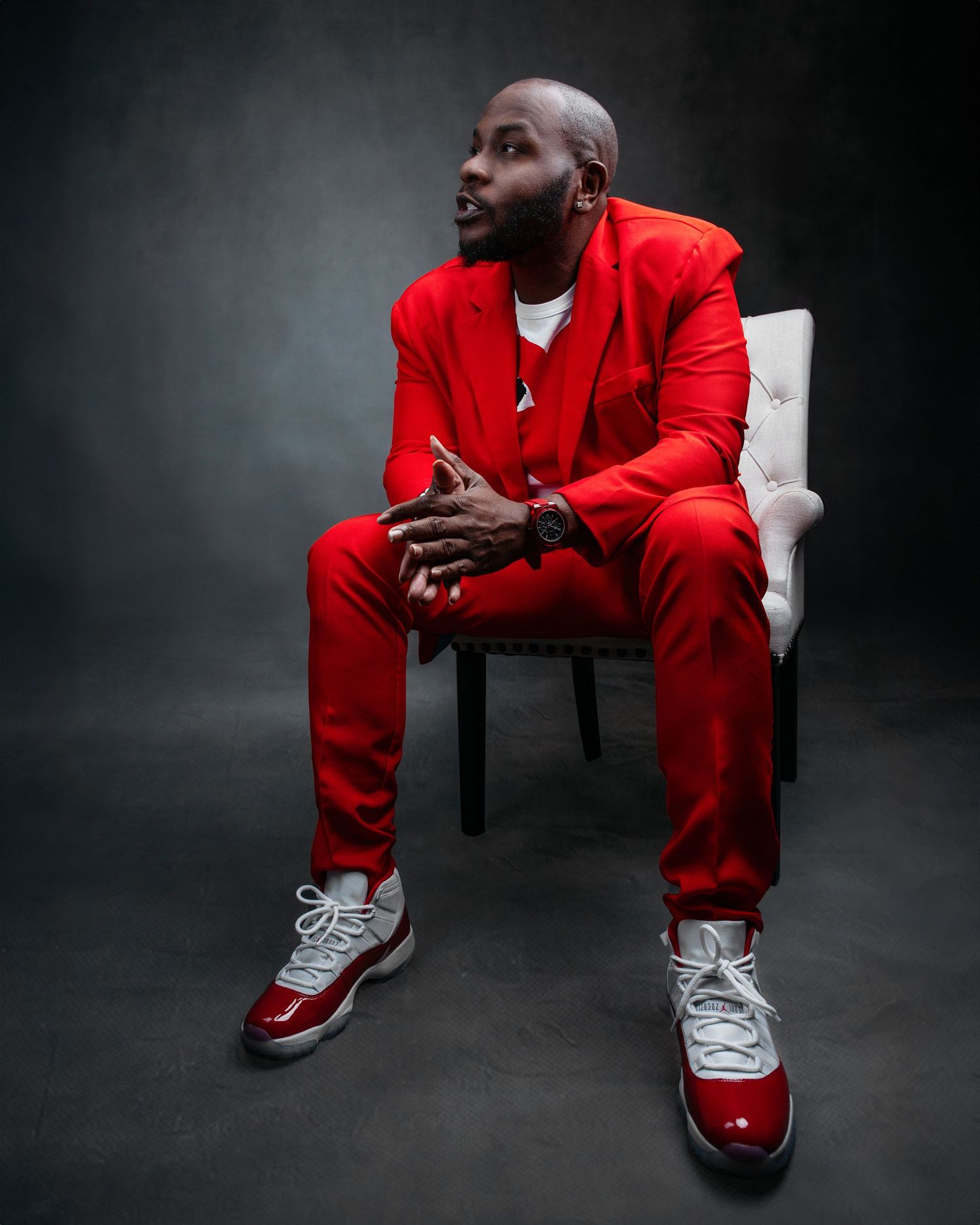Person in bright red outfit and white sneakers seated on chair against dark background.