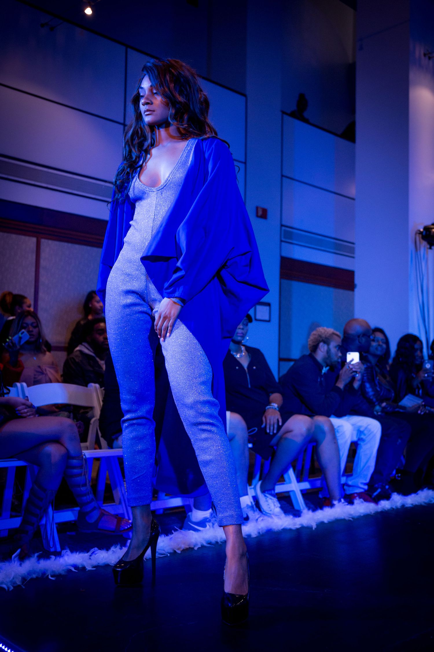 Model walks runway in bright blue coordinated outfit with white top during fashion show under dramatic lighting.