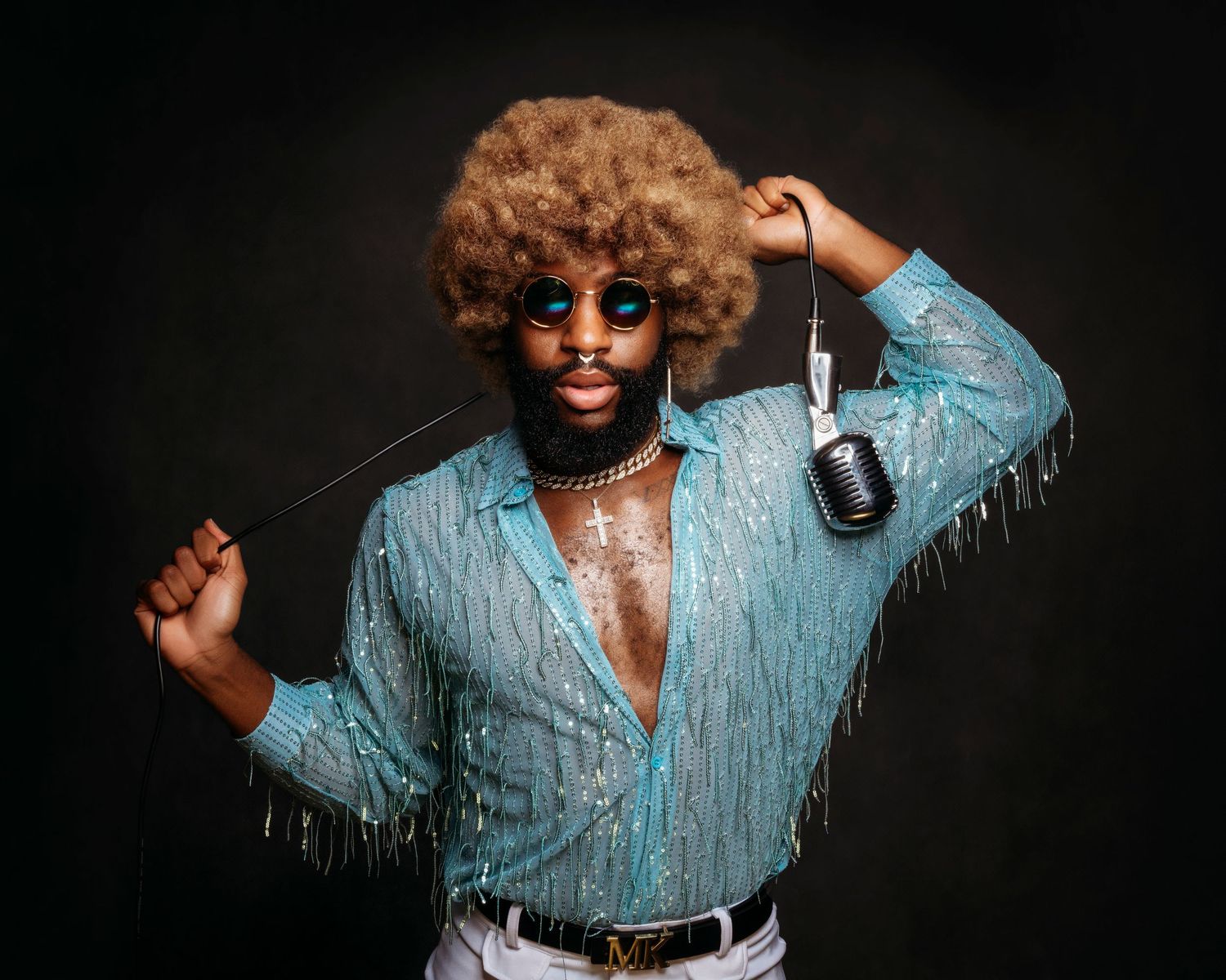Retro-styled performer in blue fringe shirt and afro hairstyle poses with microphone against dark background.