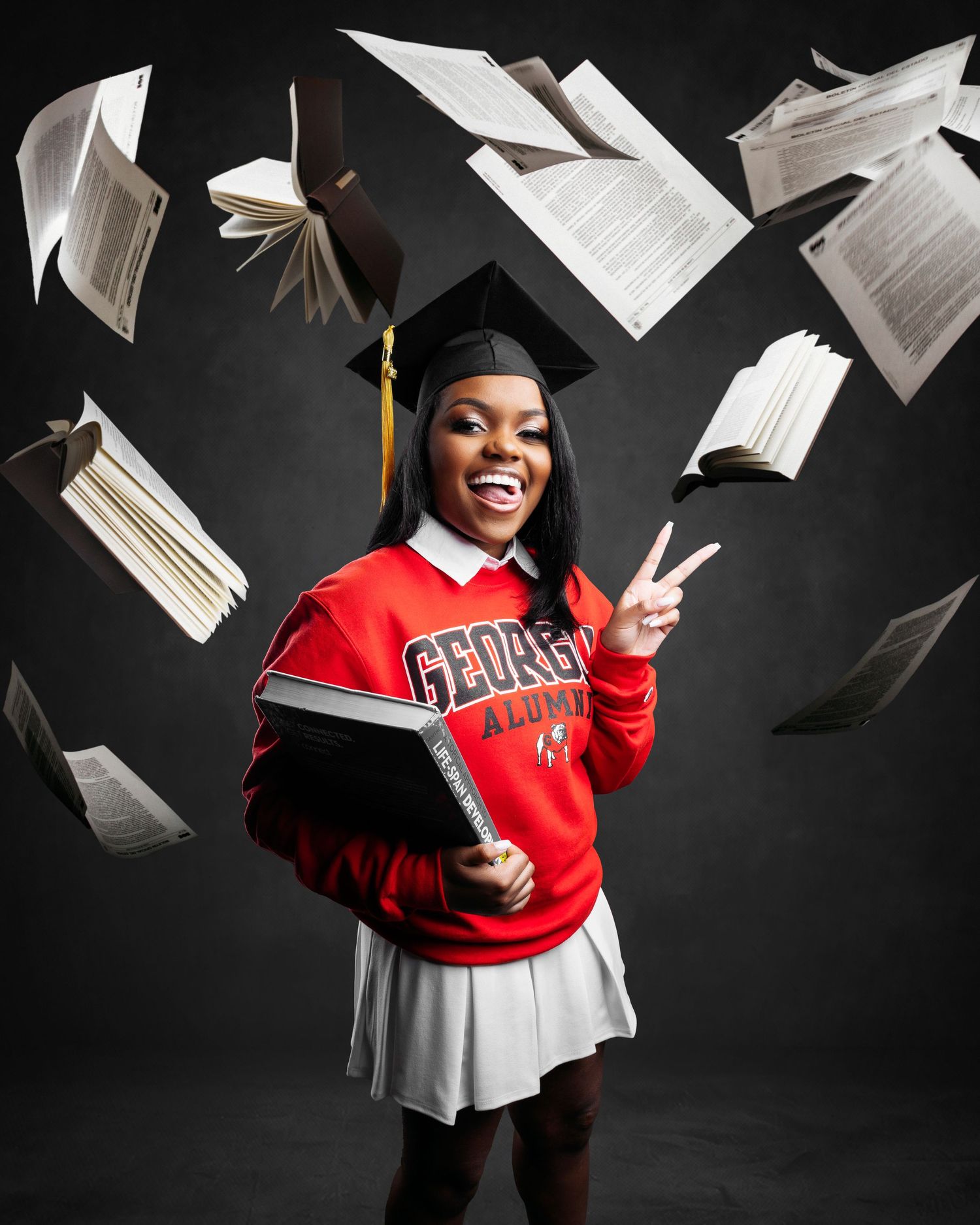 Graduation photoshoot with flying papers and red university sweatshirt against dark background.