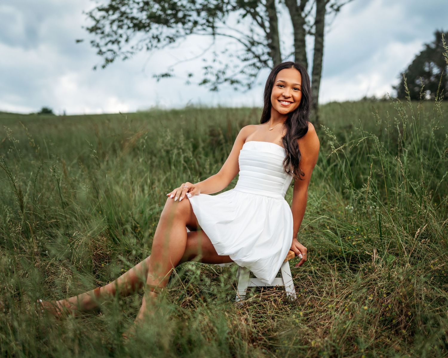 Someone in a white dress sits casually on a chair outdoors among tall grass.