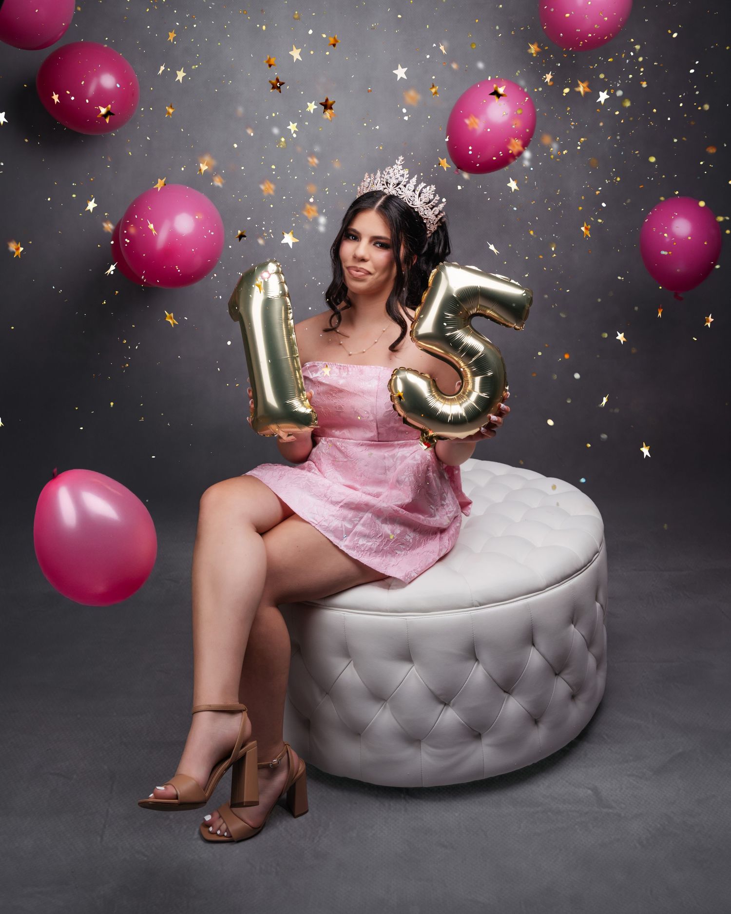 Celebratory portrait with pink balloons and confetti against grey background on tufted ottoman.