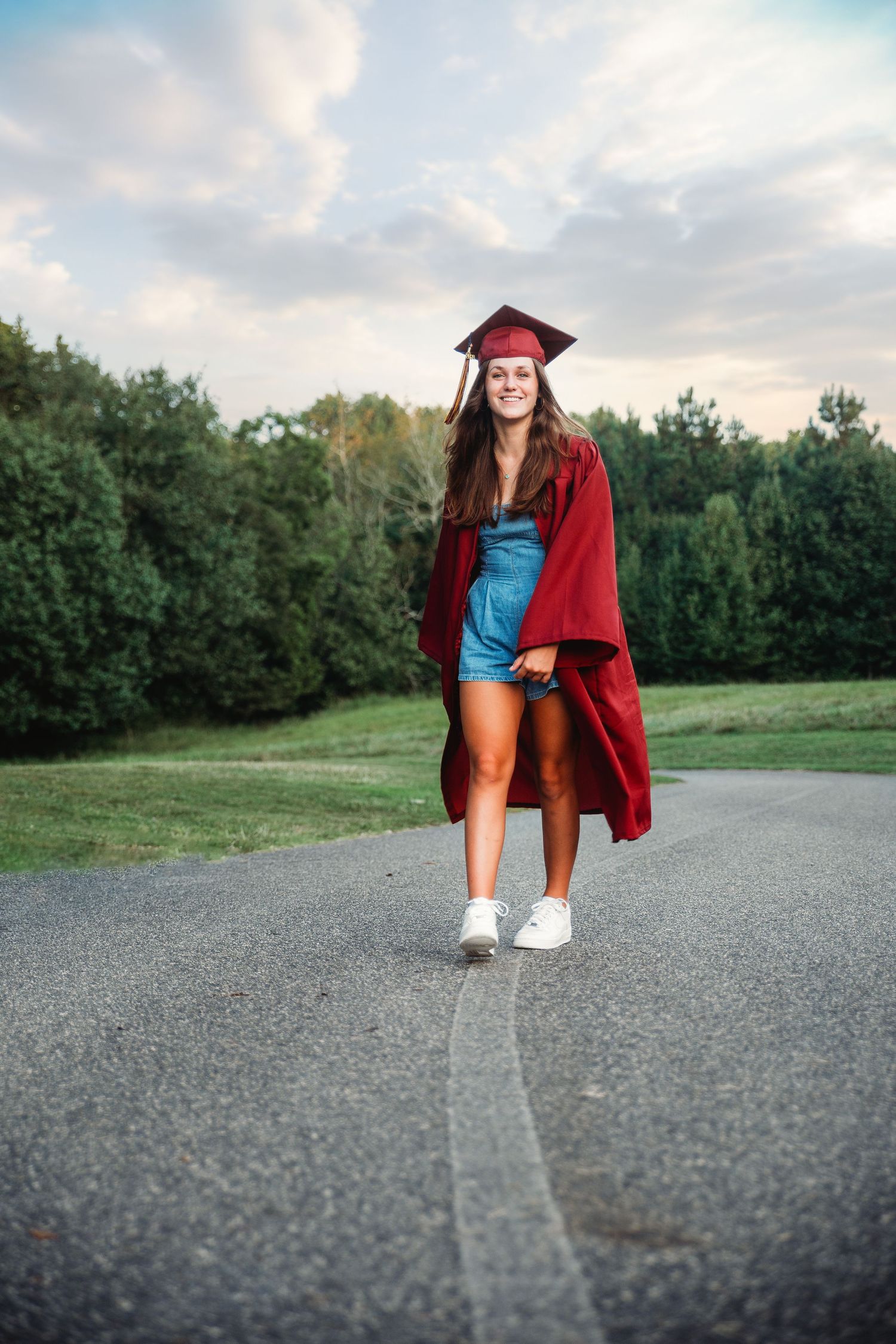 Graduate in red cap and gown poses on outdoor path during sunset celebration photoshoot.