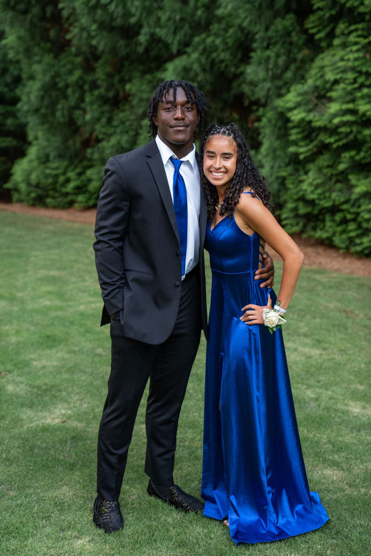 Couple in formal attire posing together outdoors, with blue dress and black suit against natural backdrop.
