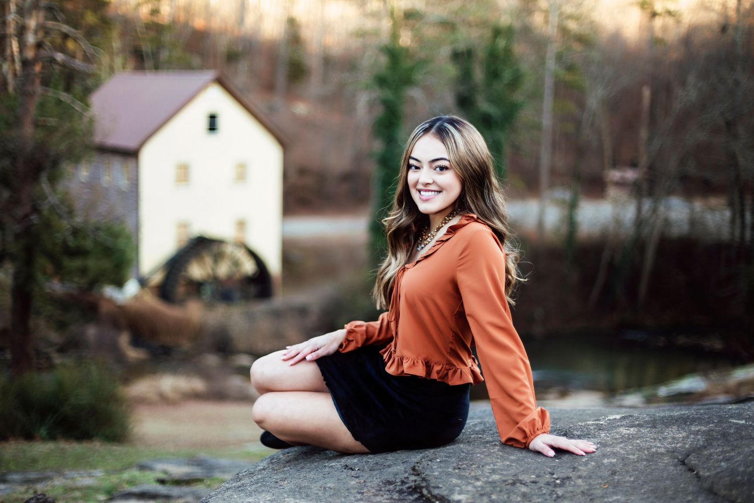 A portrait of someone in an orange top sitting by a scenic mill pond at sunset.
