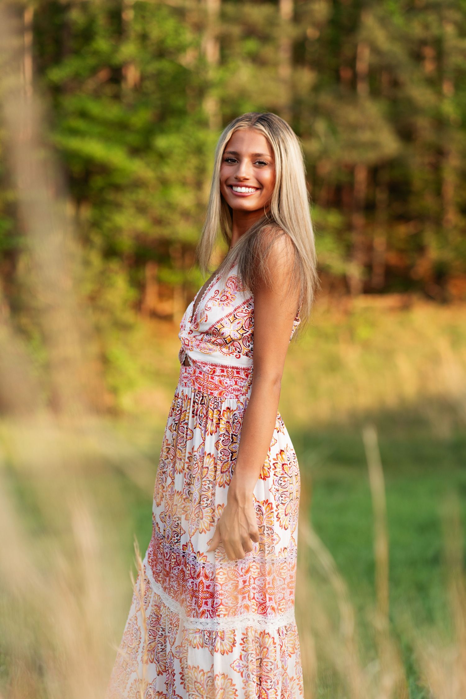 A summer portrait of someone in a patterned maxi dress standing in a sunlit forest clearing.