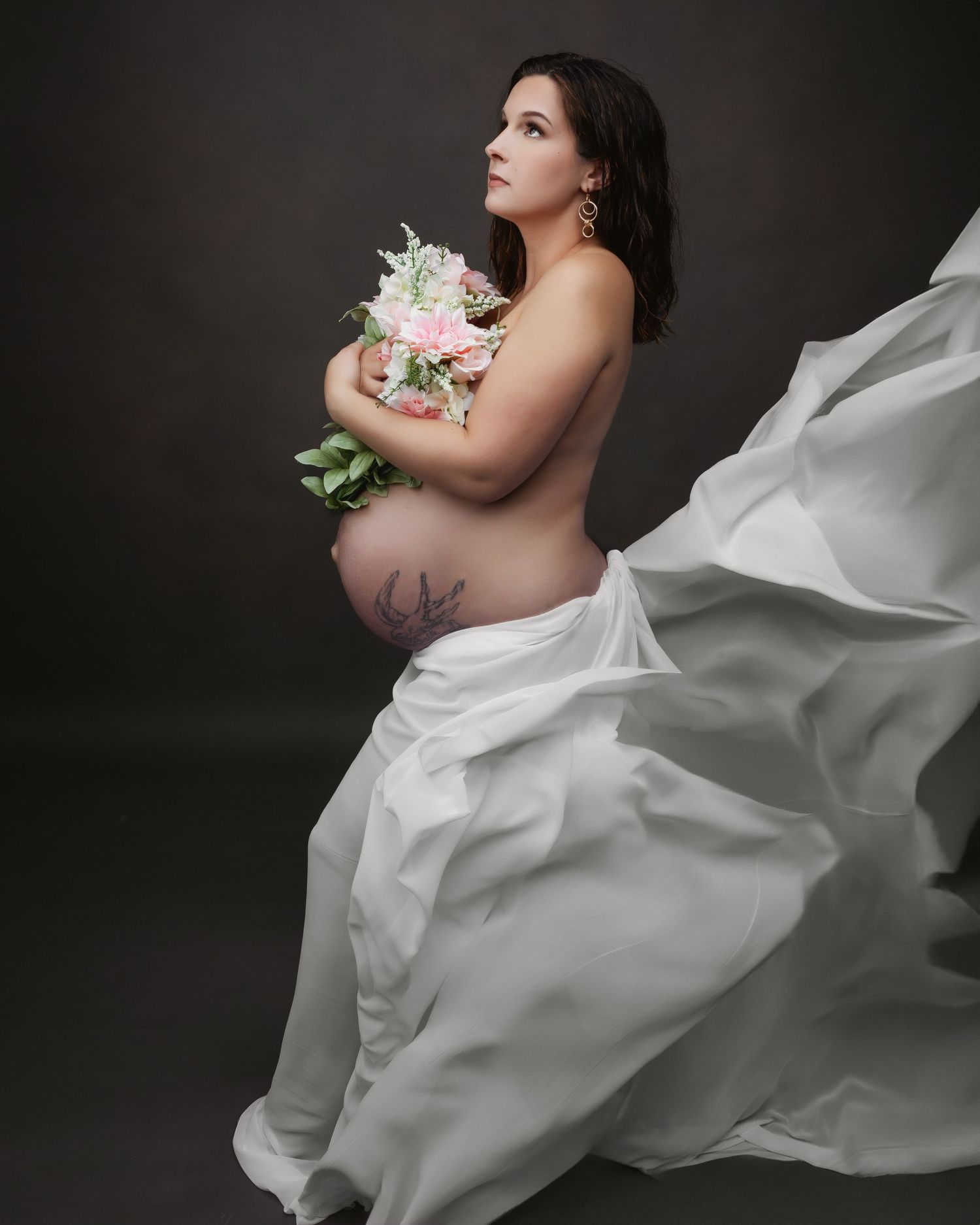Artistic maternity photo with flowing white fabric and flowers against dark background.