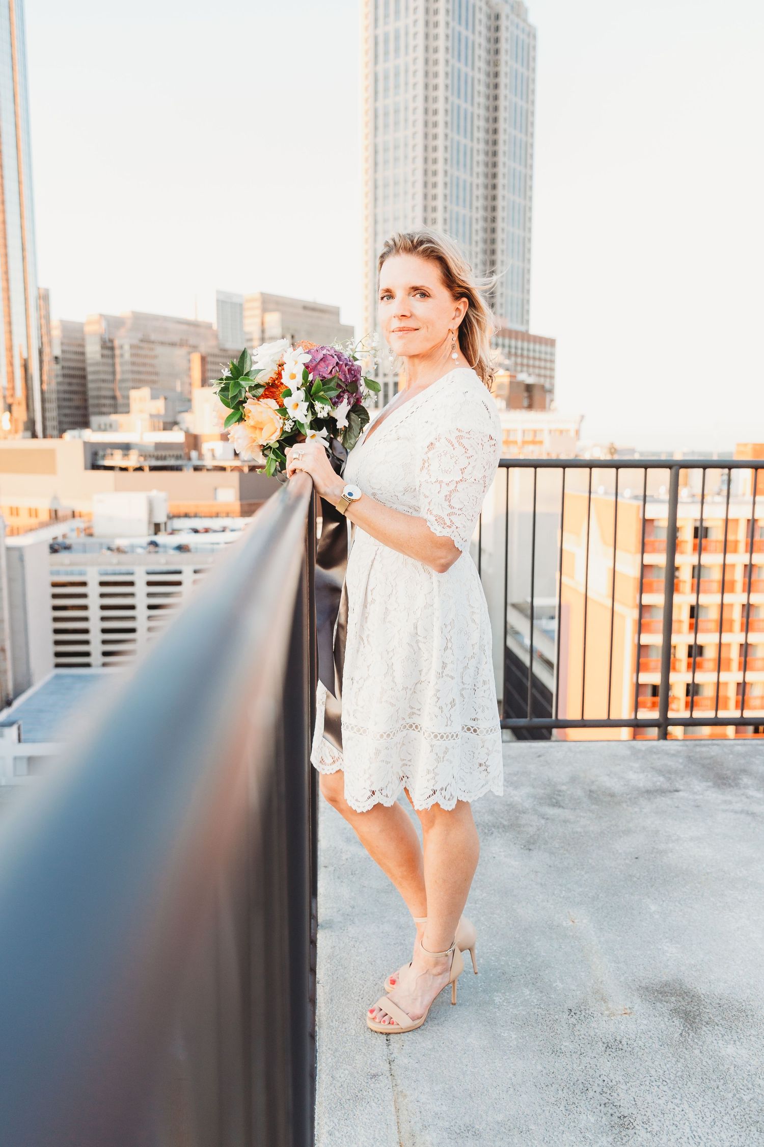 Bride in white lace dress posing on rooftop with urban skyline view.